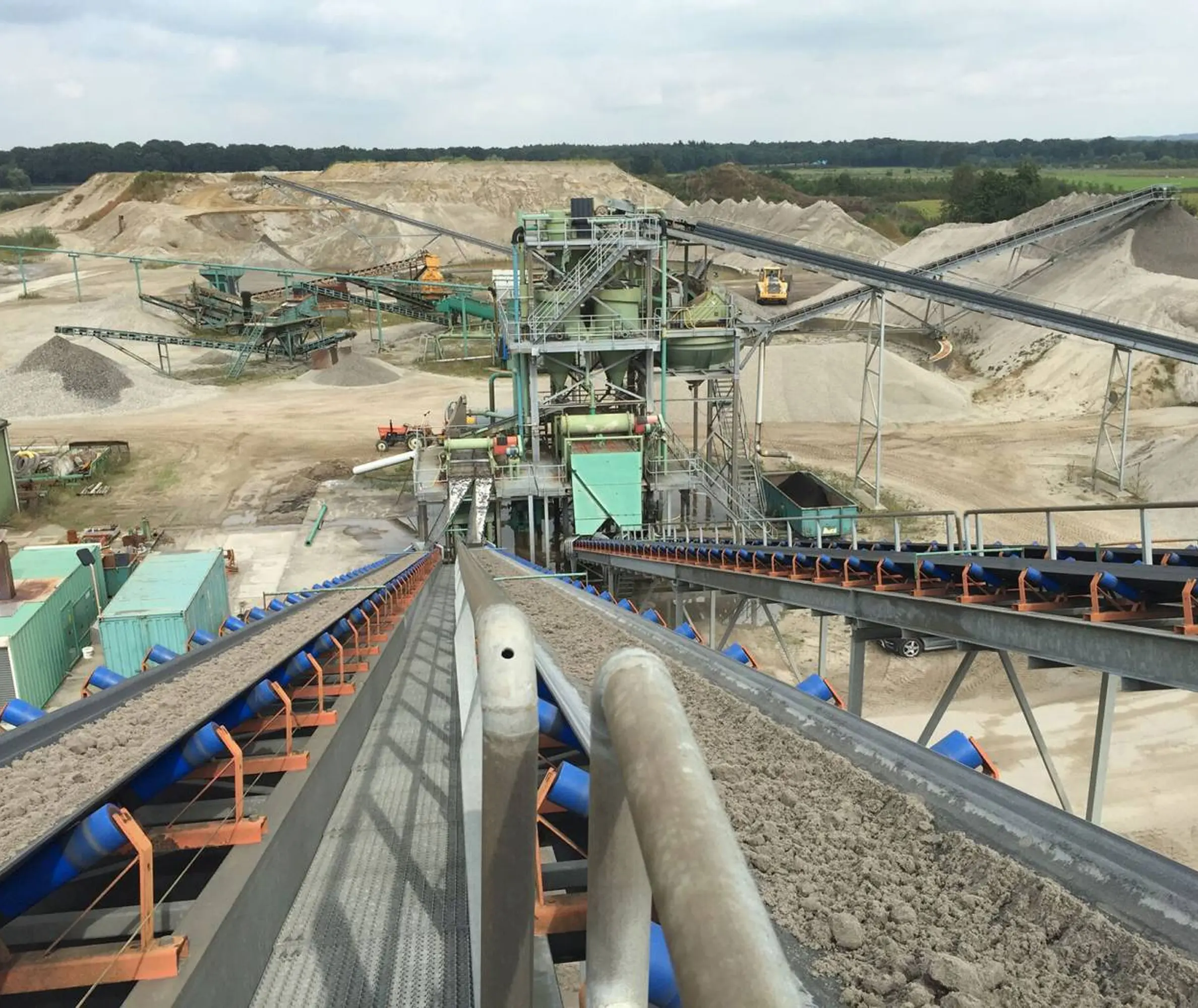 Luchtfoto van een industriële groeve met grote transportbanden die zand verplaatsen. Er zijn zandhopen en machines zichtbaar, onder een bewolkte lucht.